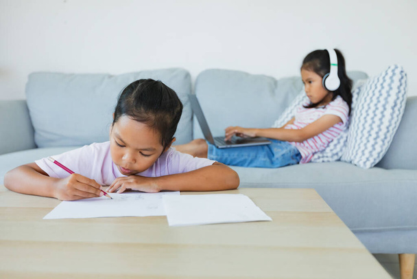 Aasian lapsi tyttö tekee laskentataulukon siskonsa kanssa istuu sohvalla ja ottaa videoneuvottelun keskustella opettajan ja luokan ryhmä. Lapset käyvät kotikoulua karanteenin aikana COVID-19-epidemian vuoksi.. - Valokuva, kuva