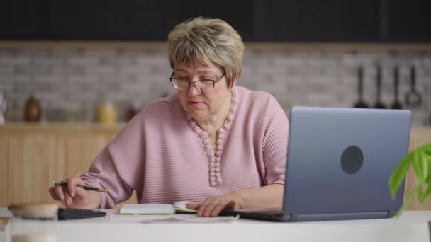 anciana está calculando facturas de servicios públicos en la cocina casera, utilizando calculadora y computadora portátil, limpieza y finanzas - Metraje, vídeo