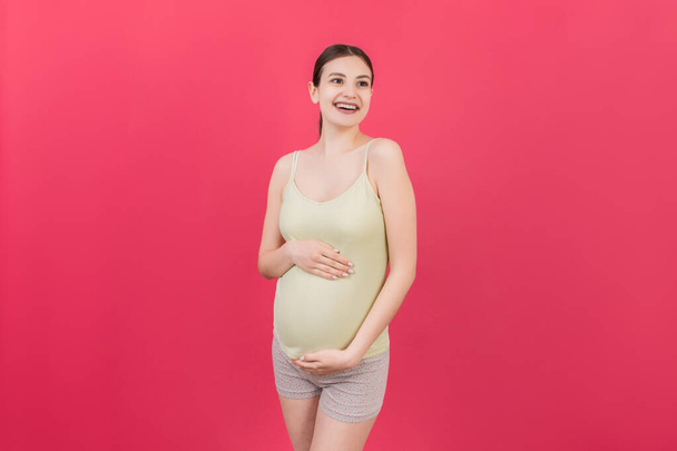 Happy pregnant woman touching her abdomen at Colored background. Future mother is wearing white underwear. Expecting of a baby. Copy space. - Fotografie, Obrázek
