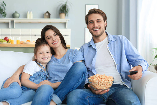 Onnellinen perhe lapsen kanssa istuu sohvalla katsomassa televisiota ja syömässä popcornia. - Valokuva, kuva