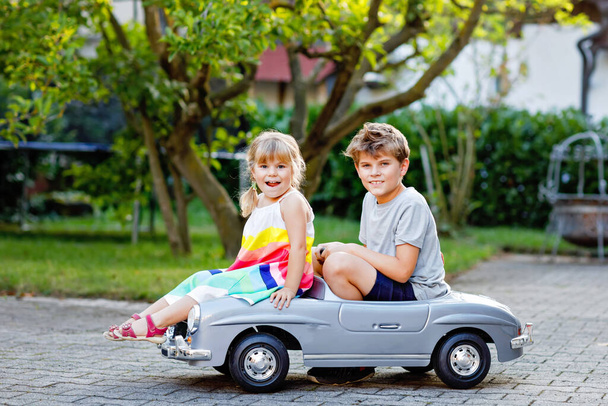 Two happy children playing with big old toy car in summer garden, outdoors. Kid boy pushing and driving car with little toddler girl, cute sister inside. Laughing and smiling kids. Lovely family - Photo, image