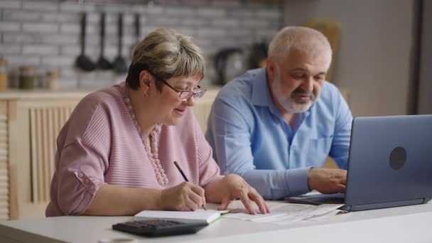 vrolijke oudere echtgenoten zitten samen in de keuken en berekenen rekeningen van nutsvoorzieningen, oude man en vrouw gebruiken online service voor het betalen van - Video