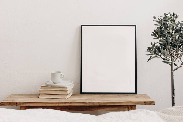 Vertical black picture frame mockup on vintage bench, table. Cup of coffee on pile of books. Potted olive tree. White wall background. Scandinavian interior, neutral color palette. Selective focus. - Foto, imagen