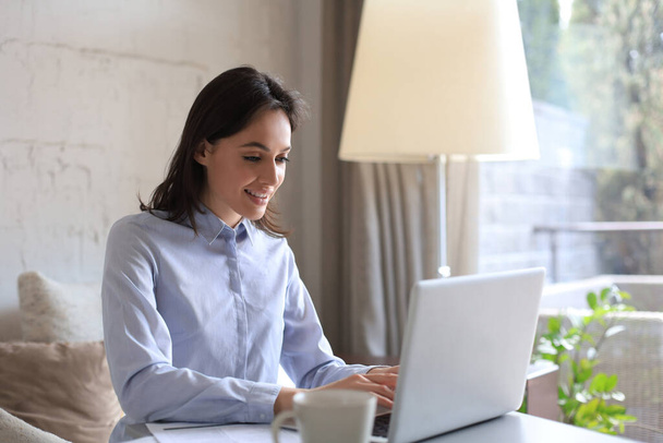 Smiling pretty woman sitting at table, looking at laptop screen. Happy entrepreneur reading message email with good news, chatting with clients online. - Photo, image