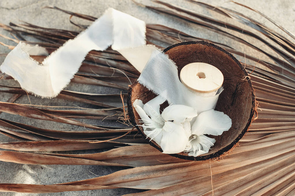Summer tropical lifestyle scene. Oleander blossoms and white silk ribbons spools in coconut shell. Dry palm leaves. Grunge concrete background in sunlight, long shadows. Top view. Selective focus. - Photo, Image