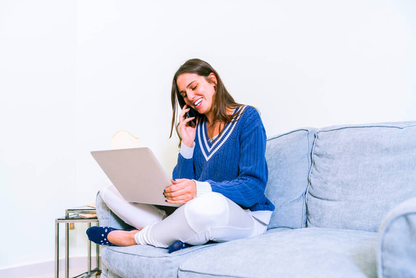 Счастливая молодая женщина сидит на диване со скрещенными ногами и использует ноутбук на сером фоне. Изображение веселой американской женщины с ноутбуком, сидящей на диване в гостиной. - Фото, изображение
