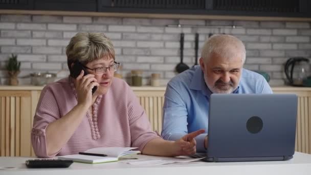 ζευγάρι συνταξιούχων είναι σύμβουλος από το τηλέφωνο, ηλικιωμένη γυναίκα καλεί και μιλάει, ο σύζυγος σερφάρει στο διαδίκτυο από το laptop - Πλάνα, βίντεο