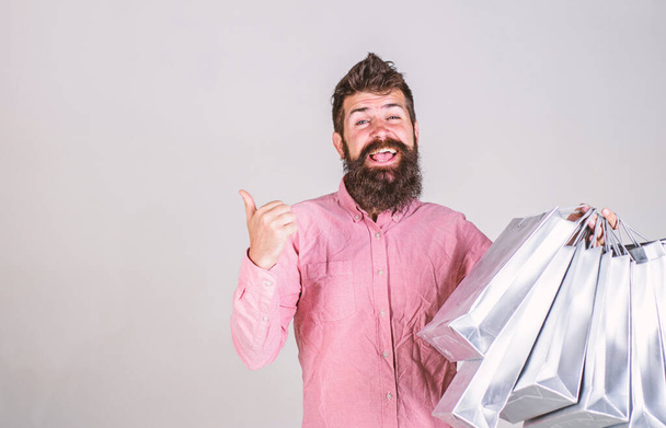 Hipster on smiling face recommends to buy. Man with beard and mustache carries bunch of shopping bags, grey background. Guy shopping on sales season, pointing at blank surface. Recommendation concept - Foto, Imagem