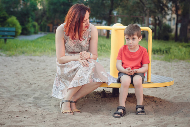 Äiti ja poika leikkikentällä. Poika kirkkaanpunaisessa t-paidassa ja nuori nainen mekossa ulkona kesällä. Jokapäiväistä elämää. Lapsuuden käsite ja lasten suhde vanhempiin - Valokuva, kuva