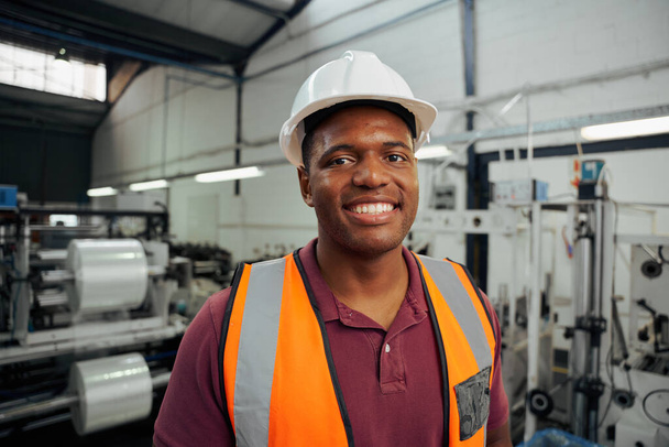 Smiling portrait of a young male factory worker wearing helmet in warehouse - Fotó, kép