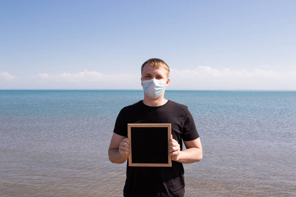 um cara com uma máscara médica azul fica no mar com um tablet vazio em suas mãos. - Foto, Imagem