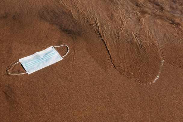 Una máscara médica azul lanzada al agua, llevada a tierra por la ola. Contaminación de la naturaleza con residuos médicos. - Foto, Imagen