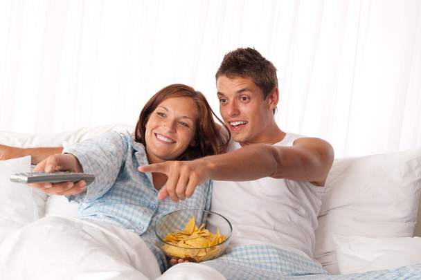 Young man and woman lying down in white bed watching television and eating crisps - Фото, зображення