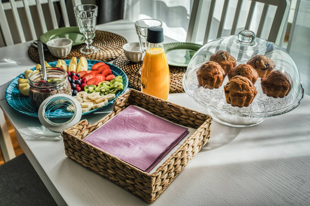 Morgens kontinentales Frühstück mit Muffins und saisonalen Früchten wie Äpfeln, Erdbeeren, Kiwis und Bananen auf weißem Holztisch mit rustikaler Strohmatte. Chia-Pudding und Beeren runden die mediterrane Bio-Mahlzeit ab, die von der Morgensonne beleuchtet wird - Foto, Bild