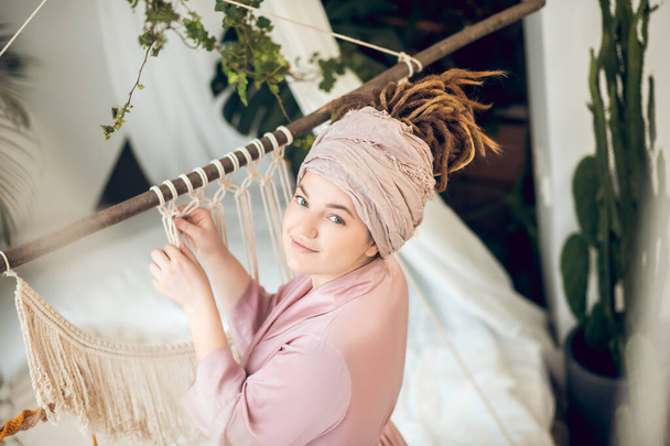 Young woman in a headwear weaving macrame and looking positive - Фото, зображення