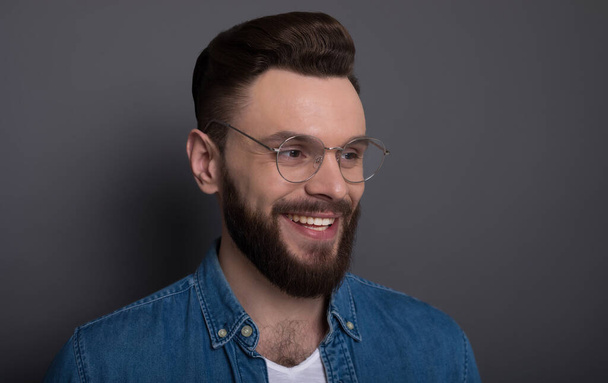 Confident smart modern bearded man in casual denim clothes and eyewear is posing over gray background in the studio - Foto, Imagen