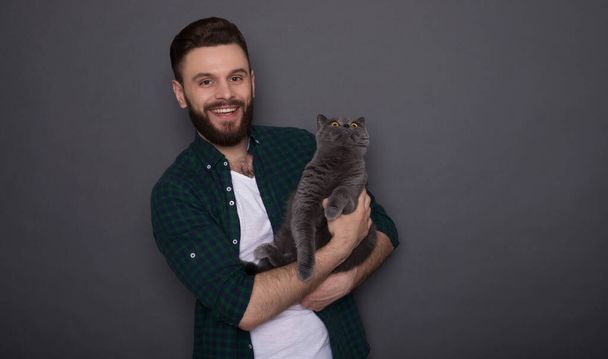 Όμορφος χαμογελαστός νεαρός γενειοφόρος άντρας κρατά την όμορφη χνουδωτή γάτα του στα χέρια και ποζάρουν μαζί σαν κολλητοί στην κάμερα. - Φωτογραφία, εικόνα