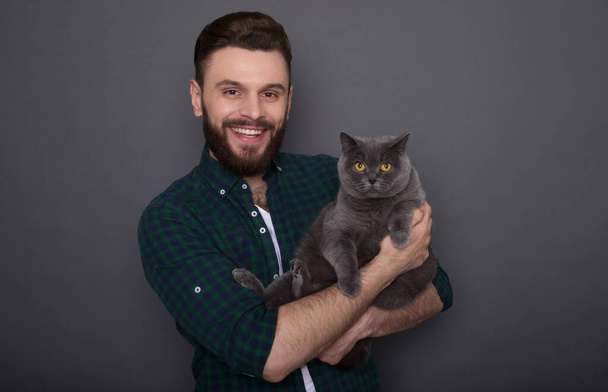Beautiful smiling young bearded man holds his lovely fluffy cat on hands and they posing together like best friends on the camera - Fotó, kép