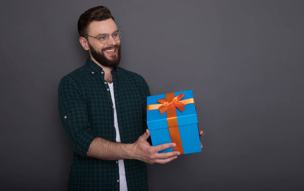 Happy young handsome bearded man in casual wear in good mood with big gift box in his hands having fun and posing over gray background - Fotografie, Obrázek