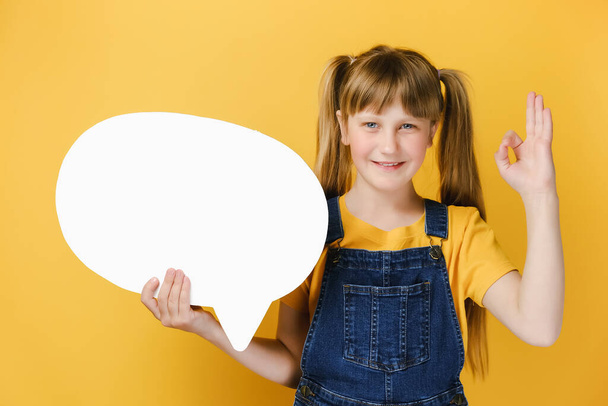 Beautiful smiling cute girl child standing over yellow studio wall background holding white speech bubble showing okay gesture. People emotion lifestyle and blogging blog website strategy concept - Valokuva, kuva