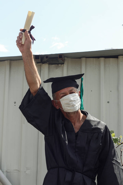 Ένας ηλικιωμένος που φορούσε καπέλο και ρόμπα αποφοίτησης με ιατρική μάσκα στο πρόσωπό του. - Φωτογραφία, εικόνα