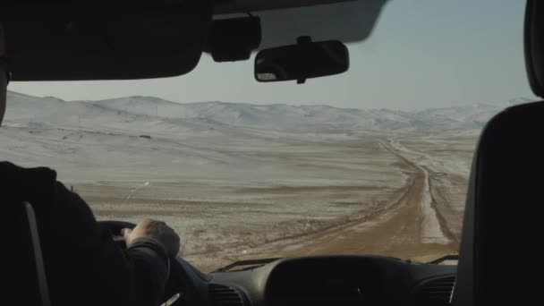 Pohled předním sklem auta na zimní silnici mezi zasněženými poli a horskými masivy u jezera Bajkal - Záběry, video