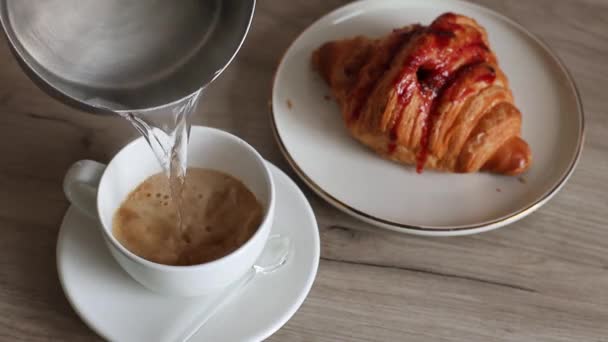 Herkullinen tuore ranskalainen croissant mansikkatäytteellä ja kupillisella aromaattista kahvia puupöydällä. Kaatamalla kuumaa vettä kuppi mustaa kahvia. - Materiaali, video