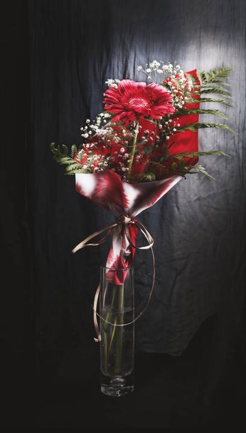 Un vase en verre avec une fleur rouge entourée de papier d'emballage sur un fond sombre - Photo, image