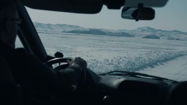 Un uomo guida una macchina su una strada invernale sullo sfondo delle catene montuose che circondano il lago ghiacciato Baikal - Filmati, video