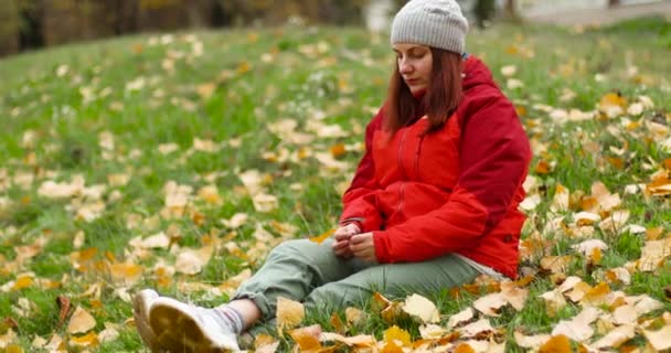 Jeune belle fille caucasienne en vêtements chauds et chapeau se trouve sur l'herbe avec des feuilles tombées dans le parc. Fille main tenant une feuille tombée - Séquence, vidéo