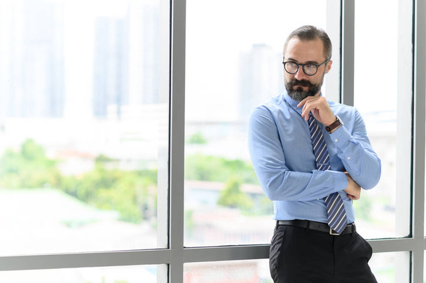 Портрет впевненого бізнесмена середнього віку в блакитній сорочці і в окулярах з бородою і вусами, що стоять поруч з вікном в офісі з копіювальним простором
 - Фото, зображення