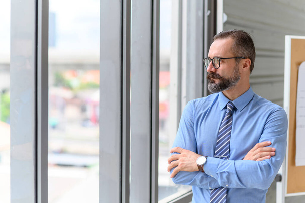 Portrait d'homme d'affaires confiant d'âge moyen en chemise bleue et portant des lunettes avec une barbe et une moustache debout à côté de la fenêtre dans le bureau avec espace de copie - Photo, image