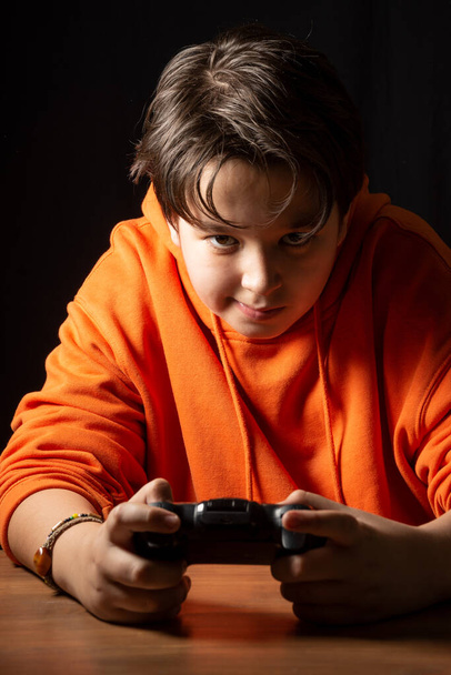 Gros plan d'un adolescent avec un sweat-shirt orange jouant avec le contrôleur sur sa console de jeu, fond sombre, vertical - Photo, image