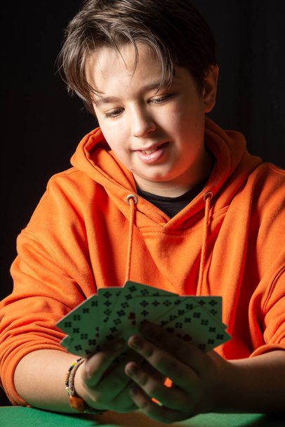 Close-up van de lachende tiener jongen, het dragen van oranje sweatshirt kijken naar zijn kaarten, donkere achtergrond, verticale - Foto, afbeelding