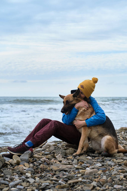 Hug the sheepdog tightly with both hands. Young cute Caucasian woman spends her free time with dog sitting on beach. Girl and a German shepherd in nature in fresh air. Love of owner and purebred dog. - Foto, Imagen