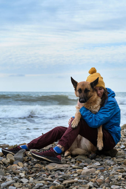 Hug the sheepdog tightly with both hands. Young cute Caucasian woman spends her free time with dog sitting on beach. Girl and a German shepherd in nature in fresh air. Love of owner and purebred dog. - Фото, зображення