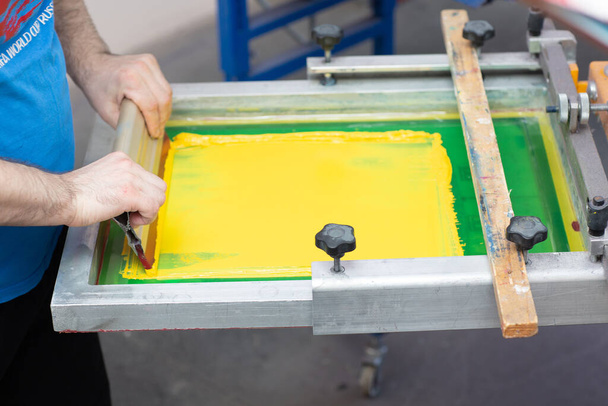 Διαδικασία εκτύπωσης μεταξοτυπίας Serigraphy στο εργοστάσιο ρούχων. Χρώματα κορνίζας, squeegee και πλαστισόλης - Φωτογραφία, εικόνα