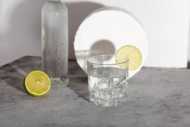 Vodka with ice and lime in glass and bottle of vodka. Shadows and sunlight trends - 写真・画像