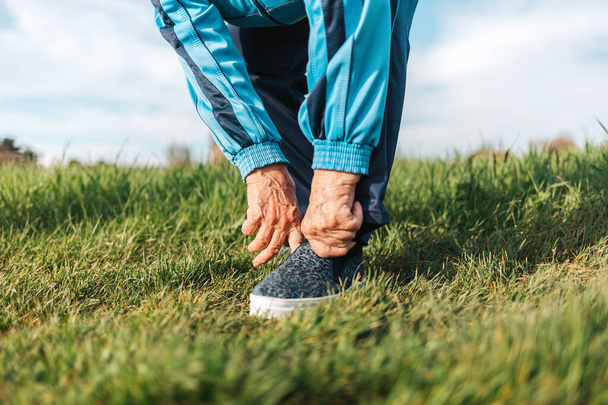 Una anciana ajusta sus zapatos deportivos en su pie mientras está de pie sobre la hierba. Primer plano. El concepto de un estilo de vida activo y problemas de salud. - Foto, imagen