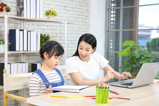 Азиатская мать работает онлайн из дома с помощью ноутбука, наблюдая за тем, как ее дочь работает над заданием художественного класса из школы для домашнего образования и концепции образования (избирательный акцент на девушке) - Фото, изображение