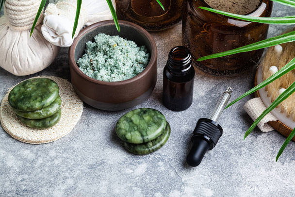 Set of natural skin care products and palm leaves on a grey background. Massage stones, essential oil, herbal compress stamps and scrub. Spa treatment concept.  - Foto, afbeelding