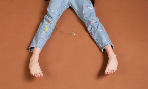 Persona irriconoscibile vestita con jeans blu e piedi nudi su uno sfondo marrone. - Foto, immagini