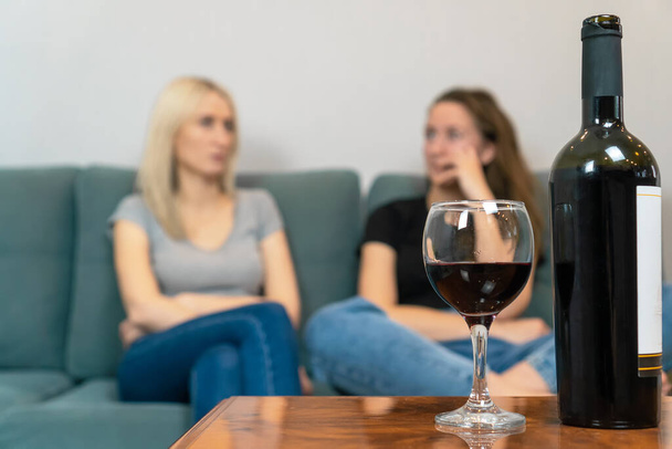 Two young women, a blonde and a brown-haired woman, are sitting on a sofa and talking, in focus in the foreground is a bottle of wine and a poured glass of wine - Photo, Image