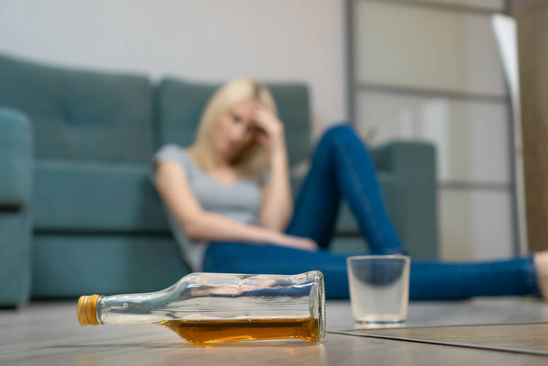 Nuori blondi nainen istuu lattialla lähellä sohvaa ja pitää päänsä, etualalla on puolityhjä pullo viskiä ja lasi. - Valokuva, kuva