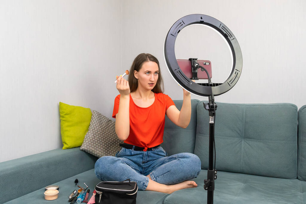 Young, cheerful, beautiful, dark-haired woman writes a beauty blog or leads a stream on a mobile phone camera with additional lighting with a ring lamp - Zdjęcie, obraz