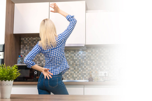 Joyeux jeune femme blonde mince en jeans bleus et une chemise cuisiniers dans la cuisine et danse, copier l'espace. Photo de derrière. Photo avec éclairage - Photo, image
