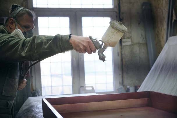 close-up Man χρησιμοποιώντας προστατευτικά γάντια ζωγραφική ξύλινη ξυλεία με πιστόλι ψεκασμού χρώμα. ψεκασμός όπλο πάρει χρώμα πάνω από την ξυλεία. - Φωτογραφία, εικόνα