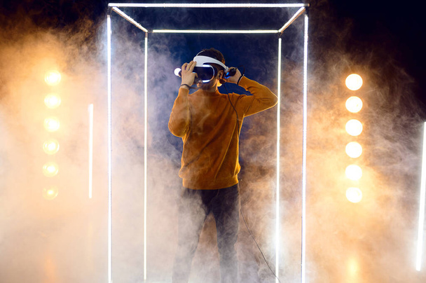 Jeune homme joue le jeu en utilisant un casque de réalité virtuelle et une manette de jeu en cube lumineux. Intérieur sombre du club de jeu, projecteur sur l'arrière-plan, technologie VR avec vision 3D - Photo, image