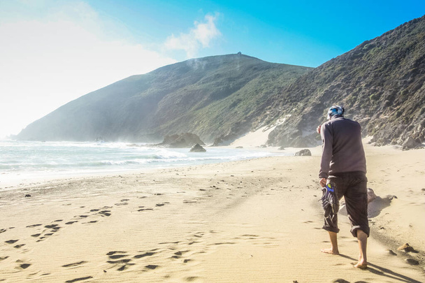 Férfi sétál a cipőjével a kezében a gyönyörű és hegyvidéki Big Sur Coast Kalifornia egy ködös, mégis napos napon, Amerikai Egyesült Államok más néven USA. - Fotó, kép