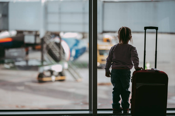 Toddler girl at airport in face mask holding a carry-on luggage. Child look at airplane after coronavirus outbreak. Safe travel and flying with child in virus pandemic. Kid at departure gate. Vacation after covid-19 lockdown. - Photo, image